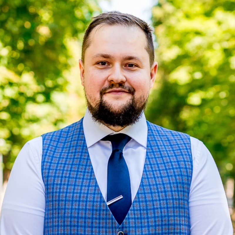 Daniel Rusu, bearded, in a checked waistcoat and dark tie, photographed outdoors under trees.